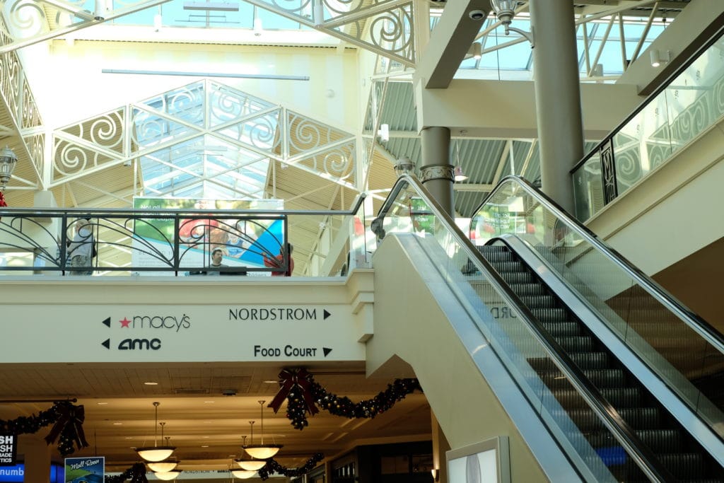 Interior of Mayfair Mall in Wauwatosa, WI with a sign pointing to different department stores, a movie theater, and food court, as well as an escalator.