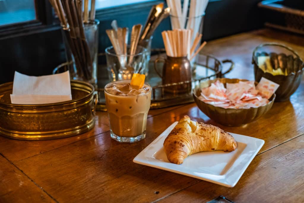 Wooden table with cutlery, a croissant on a plate, and a coffee beverage.